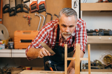 Senior carpenter uses a ruler and set square to measure and assemble a wooden structure with precise alignment, focusing on craftsmanship. Workshop is organized with tools and materials