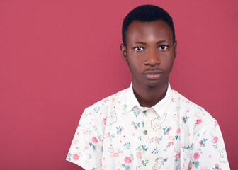 Close-up headshot of a young man with short black hair, wearing a floral shirt, making a neutral expression. His calm and composed face is set against a coral background in a studio portrait.