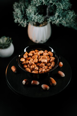 Hazelnuts arranged in a black plate surrounded by nuts on a dark surface with greenery in the background