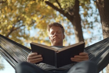 Fototapeta premium Caucasian mature male relaxing in hammock reading book outdoors