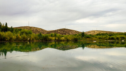 Summer landscape calm river, lake reflecting mountains, green trees against cloudy sky.
