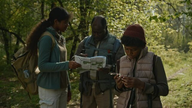 Teenage boy navigating with compass while his elder sister and father reading map in woods during autumn hike