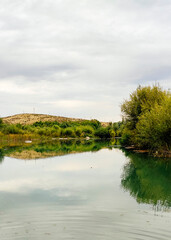 Summer landscape calm river, lake reflecting mountains, green trees against cloudy sky.
