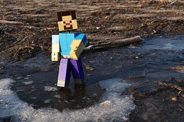 Naklejka premium LEGO Minecraft action figure of Steve walking on frozen water puddle during freezing winter morning, sunlit by daylight sunshine. 