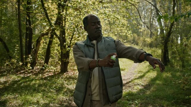 African American mature male tourist applying mosquito repellent on his shirt and vest when hiking on forest trail in early fall