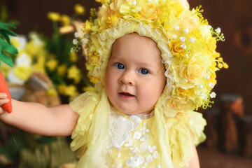 Portrait of a little girl in a yellow suit. Girl 7 months old sitting. Little child in a hat made of flowers. Little child holding a carrot in his hands