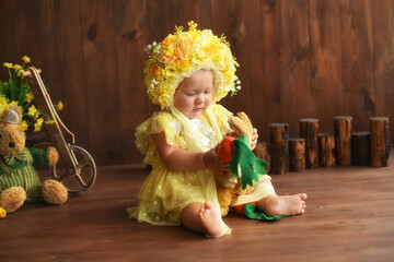 Portrait of a little girl in a yellow suit. Girl 7 months old sitting. Little child in a hat made of flowers. Little child holding a carrot in his hands