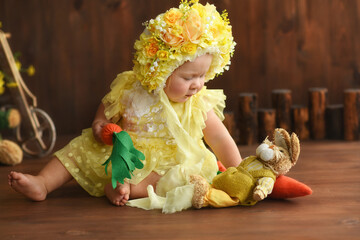 Portrait of a little girl in a yellow suit. Girl 7 months old sitting. Little child in a hat made of flowers. Little child holding a carrot in his hands