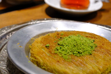 Close-up of traditional Turkish syrupy dessert kunefe. Kunefe with pistachios. Kadayif desserts. Lemon and brewed tea in a glass cup. Presentation on a copper plate. Turkish desserts. Food industry.