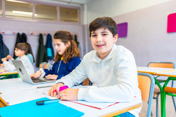 Elementary school students studying in classroom, using laptop and colorful markers