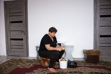 Middle-aged woman is sitting on a stool peeling potatoes next to a white bucket inside a room with two doors. The scene is set on an ornamental carpet, evoking a domestic and traditional feel.