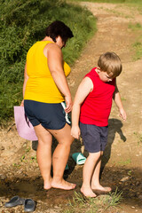 Fototapeta premium A woman and young boy playfully walk through a shallow stream. They are wearing casual summer clothing and appear to be enjoying a relaxing day in nature.