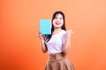 Naklejka premium A woman wearing a white shirt and a brown skirt holding a blue book points to the side on orange background