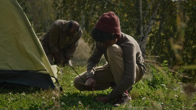 Teenage boy and his dad pulling guy ropes and staking them into ground while setting up rainfly on their tent during autumn hike