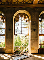 Abandoned mansion interior with broken arched windows, torn curtains, and scattered debris. Sunlight illuminates decayed walls, shattered glass, and a collapsed balcony railing, creating an eerie yet