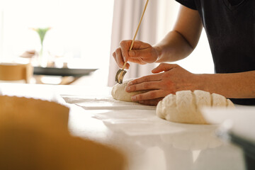 Woman hands skillfully score the surface of a round dough loaf, preparing sourdough bread for baking. The bright kitchen setting provides a warm atmosphere and fresh ingredients.