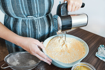 Woman cook in an apron mixes dough in plastic bowl with mixer to prepare a cake on kitchen table.