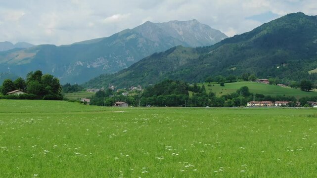 Clusone plateau, drone flying over green fields and meadows with mountains in background, Rovetta, Italy. Aerial forward at low altitude