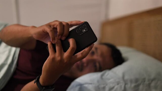 Asian man lying on the bed while using smartphone befor sleeping. Selective focus on phone
