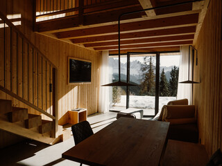 A modern wooden chalet living space bathed in natural light, with expansive windows showcasing a snowy mountain landscape. A perfect blend of warmth, elegance, and nature.
