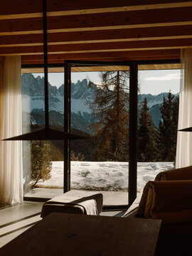 A modern wooden chalet living space bathed in natural light, with expansive windows showcasing a snowy mountain landscape. A perfect blend of warmth, elegance, and nature.