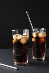 Refreshing cola with ice cubes in glasses and drinking straws on black table, closeup