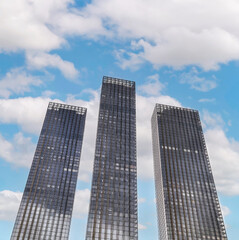 Obraz premium Three modern skyscrapers glisten under a bright blue sky with fluffy clouds during daytime in a bustling urban area
