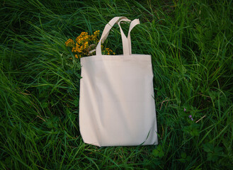 White canvas tote bag mockup template with long handles and wild flowers among the green plant leaves outdoor