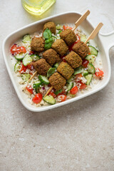 Falafel skewers with couscous and vegetables in a serving tray, vertical shot on a beige stone background with space, elevated view