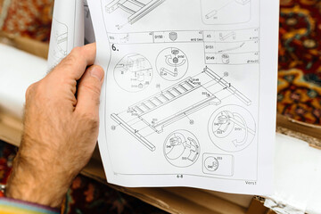A close-up of a hand holding an instruction manual with technical drawings and assembly steps for a toddler bed, featuring labeled components and screw placements.