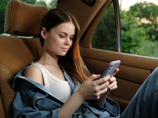 Young woman in casual denim attire using a smartphone in a car, showing a relaxed expression while...