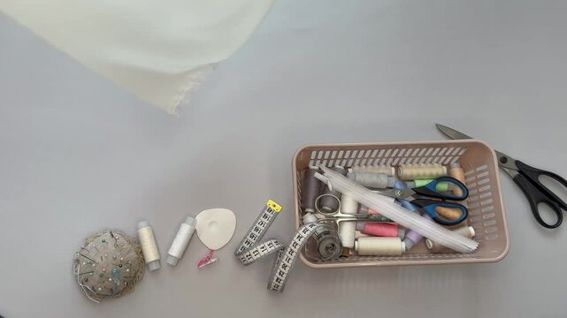 Video of white silk fabric falling on cutting table. Abov view of sewing accessories, needles, box of threads, scissors, zip, pincushion and tape measure. Sewing as hobby. Top view, closeup, flat lay 
