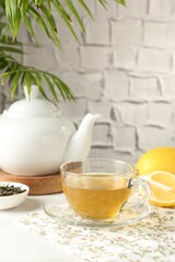 Refreshing green tea in cup, lemon and dry leaves on white table against grey textured wall