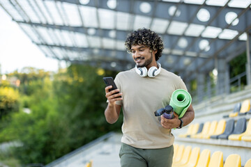 A man with a phone and a yoga mat walks in the park, an athlete uses an application on a smartphone for joint training and acquaintances, books a place in a training class.