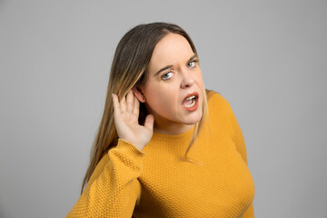 Curious blond woman trying to hear holding hand to ear on gray background