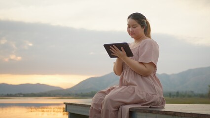 Naklejka premium Woman Using Tablet by Lake at Sunset, Surrounded by Majestic Mountains. Digital nomad freelancer, Remote work outdoors in vacation time. Work everywhere. Asian Businesswoman casual looks
