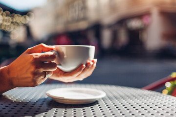 Close up of cup of coffee in female hands. Woman drinking hot beverage in outdoor cafe at sunset. Space