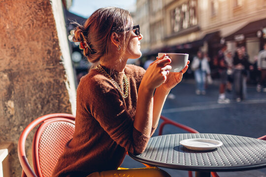 Young stylish woman sits on cafe terrace outdoor drinking coffee. Woman enjoys architecture of European city on street