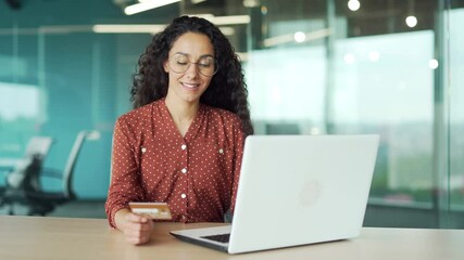 Happy young businesswoman doing online shopping typing credit card data on laptop while sitting at a workplace in business office. Smiling female employee makes a purchase, enjoy e-commerce e-shopping