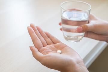 Sick asian young woman, girl holding tablet pill on hand, painkiller medicine from fever, stomach pain, headache for treatment, take drug or vitamin with glass of water at home. Health care concept.