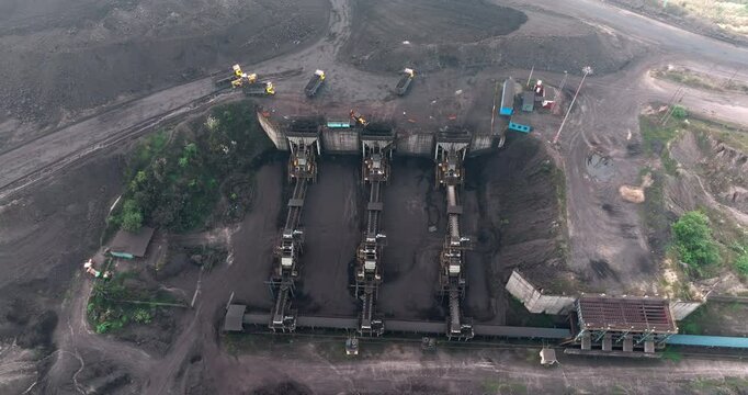 A top-angle drone shot captures trucks full of coal unloading at the crusher after arriving from the coal mine, while additional loaded trucks steadily arrive at the site.