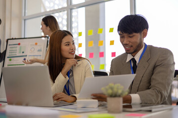 A group of people are working together in a conference room. A man and a woman are looking at a presentation on a laptop. There are several other people in the room
