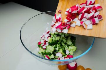 Close up of adding sliced cucumbers to green lettuce leaves in salad bowl using wooden cutting...