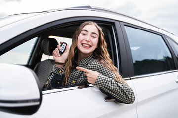 A young woman looking out of driver's seat window holding a key of new modern car. A cheerful...