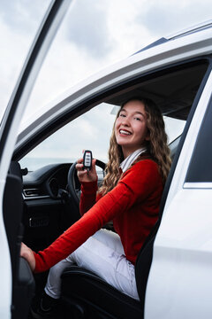 Young smiling overjoyed woman in casual wear holding a key to her new car. Happy female owner of new automobile showing a key and looking at camera. Outside of car view. Buy or rent auto. Vertical.