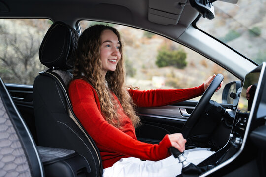 A young white woman holding steering wheel and gear shift, changing speed in the modern car. Side view portrait of a female driver looking at road shifting gears on the transmission lever.
