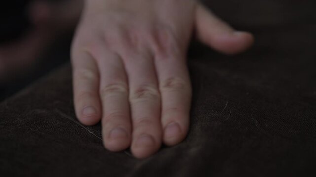 Close-up hand of unrecognizable female pet owner meticulously picking and removing cat hair from surface of dark brown sofa. Concept of common challenge of pet hair on furniture, slow motion.
