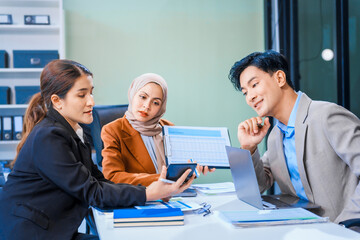Obraz premium An Asian middle-aged man, a middle-aged Muslim woman in hijab, middle-aged Asian woman collaborate during business meeting. They sit at desk, reviewing report documents and discussing teamwork.