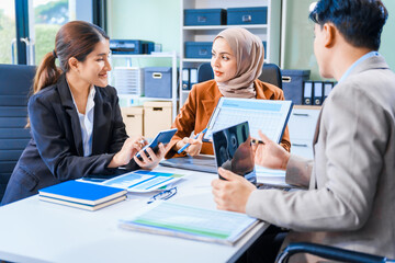 An Asian middle-aged man, a middle-aged Muslim woman in hijab, middle-aged Asian woman collaborate during business meeting. They sit at desk, reviewing report documents and discussing teamwork.