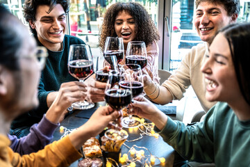 Group of friends toasting red wine on happy hour sitting at bar restaurant table - Millennial people enjoying dinner party together - Life style concept with guys and girls hanging out on weekend
